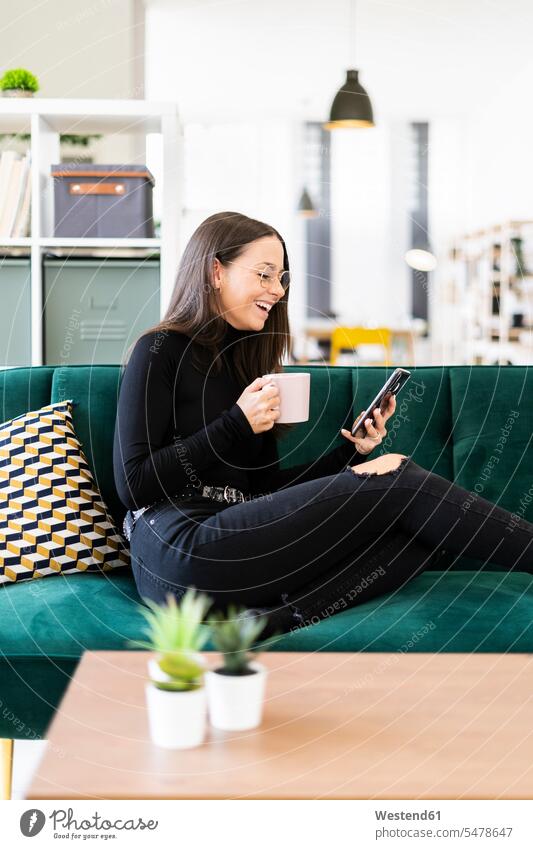 Fröhliche weibliche Bloggerin telefoniert, während sie zu Hause mit Kaffeetasse auf dem Sofa sitzt Farbaufnahme Farbe Farbfoto Farbphoto Innenaufnahme