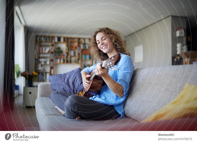 Lächelnde Frau spielt Gitarre, während sie zu Hause auf dem Sofa sitzt Farbaufnahme Farbe Farbfoto Farbphoto Innenaufnahme Innenaufnahmen innen drinnen Tag