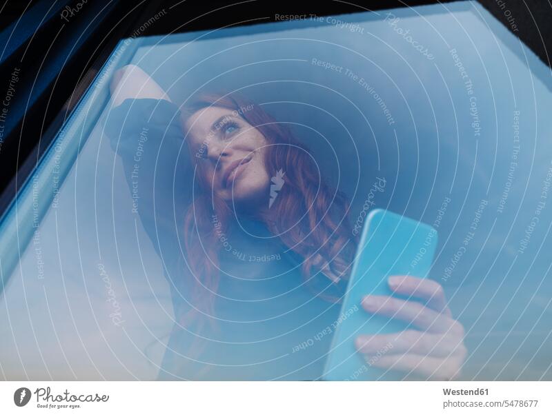 Porträt einer lächelnden rothaarigen Frau, die ihr Handy hinter einer Fensterscheibe hält geschäftlich Geschäftsleben Geschäftswelt Geschäftsperson