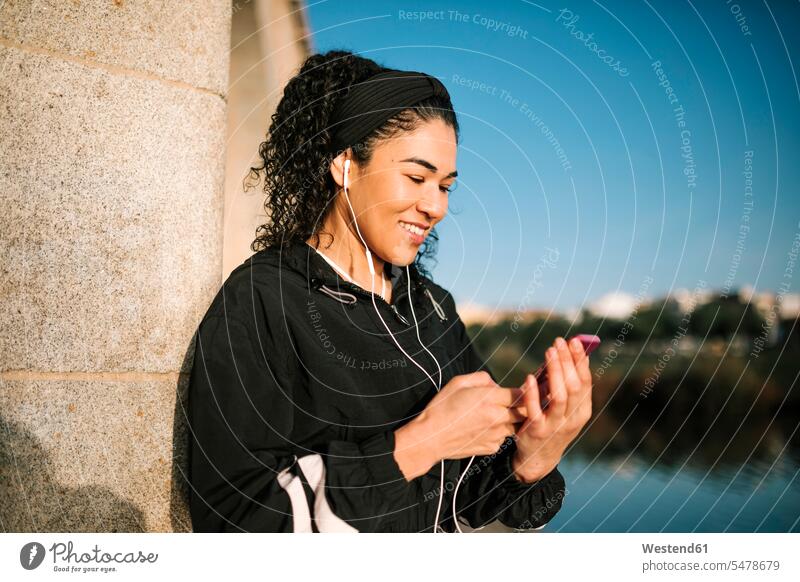 Lächelnde Sportlerin mit Mobiltelefon Musik hören, während lehnt sich auf Spalte während sonnigen Tag Farbaufnahme Farbe Farbfoto Farbphoto Außenaufnahme außen