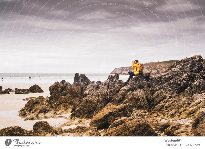 Junge Frau mit gelber Regenjacke am Strand, Blick durch ein Fernglas, Bretagne, Frankreich Rucksäcke Feldstecher Fernglaeser Ferngläser entdecken sitzend sitzt