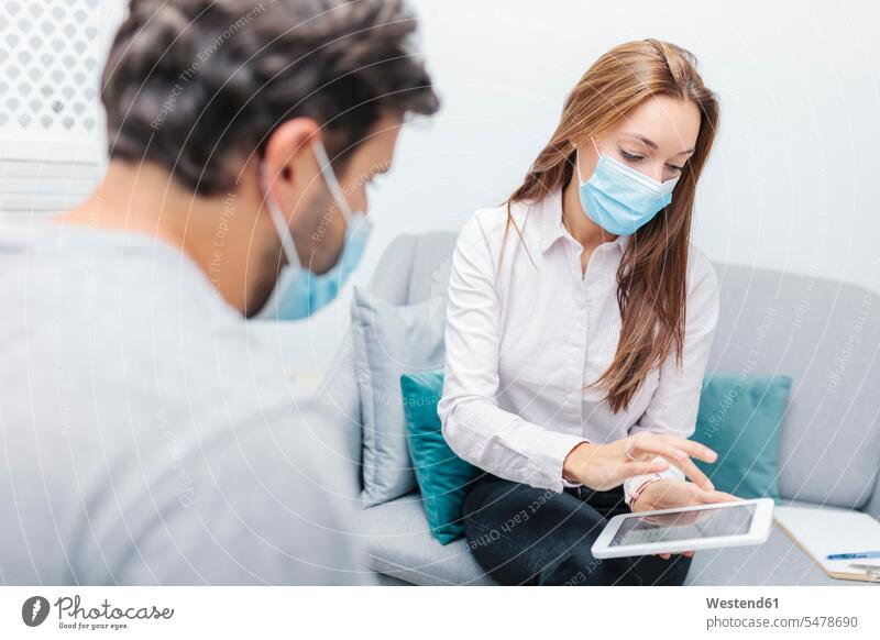 Arzt trägt Gesichtsmaske mit digitalem Tablett, während er bei einem Mann im Büro sitzt Farbaufnahme Farbe Farbfoto Farbphoto Innenaufnahme Innenaufnahmen innen