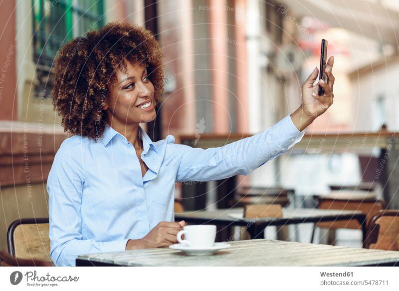 Lächelnde Frau mit Afrofrisur sitzt in einem Café im Freien und macht ein Selfie Selfies weiblich Frauen sitzen sitzend lächeln Cafe Kaffeehaus Bistro Cafes