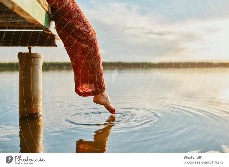 Frau sitzt bei Sonnenuntergang auf einem Steg an einem See und berührt mit dem Fuß das Wasser Leute Menschen People Person Personen Europäisch Kaukasier