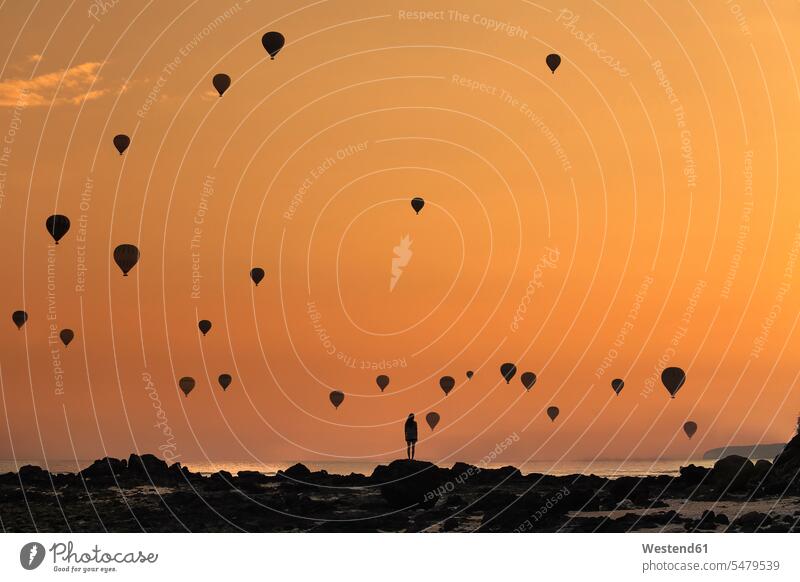 Indonesien, West-Nusa Tenggara, Silhouetten von Heissluftballons, die in der stimmungsvollen Abenddämmerung über eine einsame Frau fliegen, die an einer felsigen Küste steht