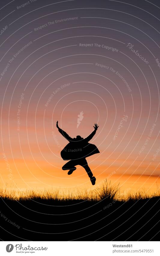 Unbekümmerter Mann mit Jacke, der mit ausgestreckten Armen gegen den Himmel bei Sonnenuntergang springt Farbaufnahme Farbe Farbfoto Farbphoto Außenaufnahme