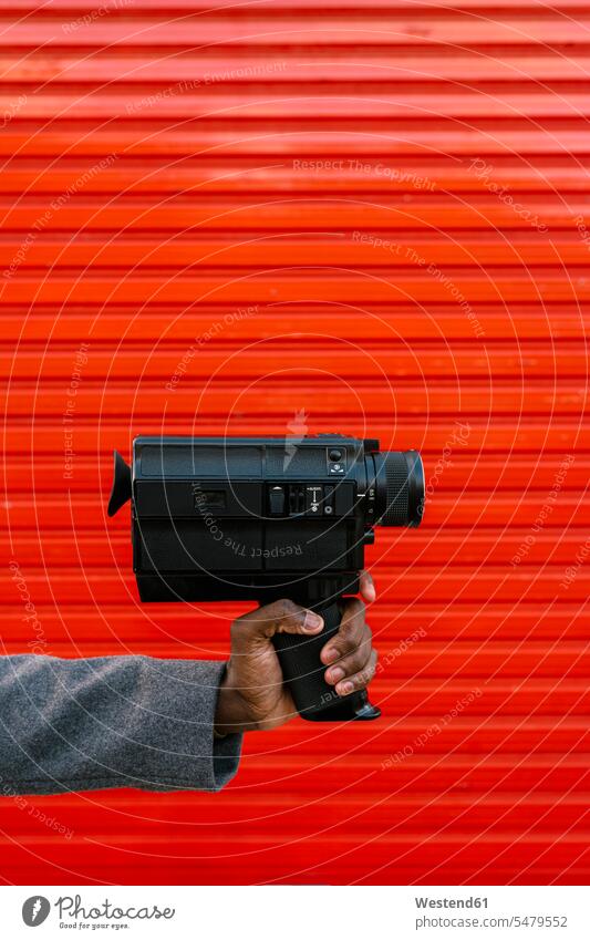 Mann hält 80er Jahre Filmkamera gegen rote Wand Farbaufnahme Farbe Farbfoto Farbphoto Außenaufnahme außen draußen im Freien Tag Tageslichtaufnahme