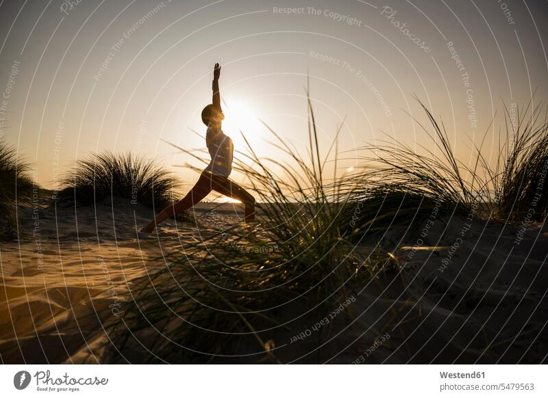 Silhouette einer jungen Frau, die bei Sonnenuntergang Yoga in Kriegerposition inmitten von Pflanzen am Strand vor klarem Himmel praktiziert Farbaufnahme Farbe