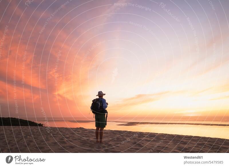 Älterer Mann mit Rucksack schaut bei Sonnenuntergang auf den Atlantik gegen den Himmel, Düne von Pilat, Nouvelle-Aquitaine, Frankreich Farbaufnahme Farbe