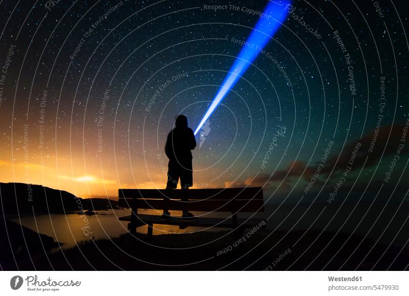 Spanien, Ortigueira, Loiba, Silhouette eines auf Bank stehenden Mannes unter Sternenhimmel mit blauem Strahl Umrisse Gegenlicht Kontur Silhouetten