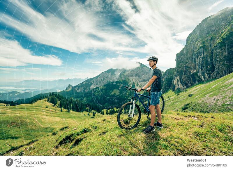 Deutschland, Bayern, Pfronten, junger Mann mit Mountainbike auf Almwiese bei Aggenstein Aussicht Ausblick Ansicht Überblick Männer männlich Mountainbikes stehen