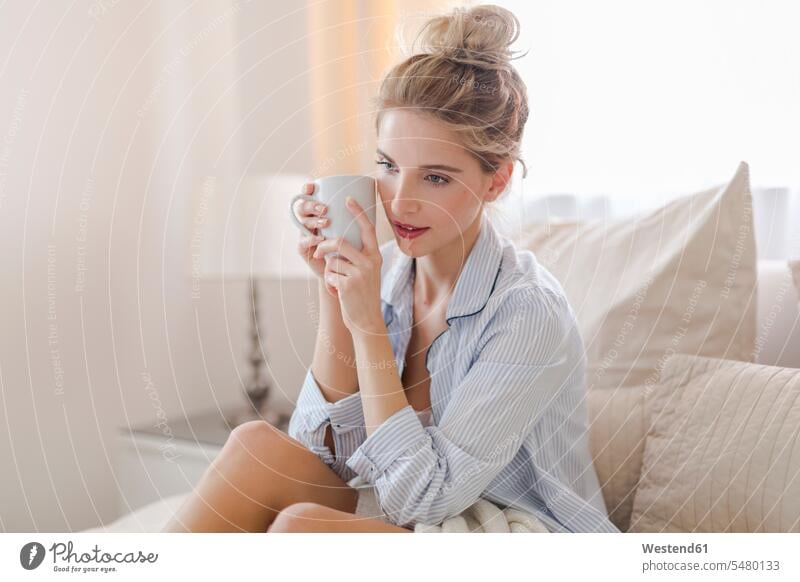 Porträt einer tagträumenden blonden Frau, die mit einer Tasse Kaffee auf dem Bett sitzt Europäer Kaukasier Europäisch kaukasisch Betten Innenaufnahme drinnen