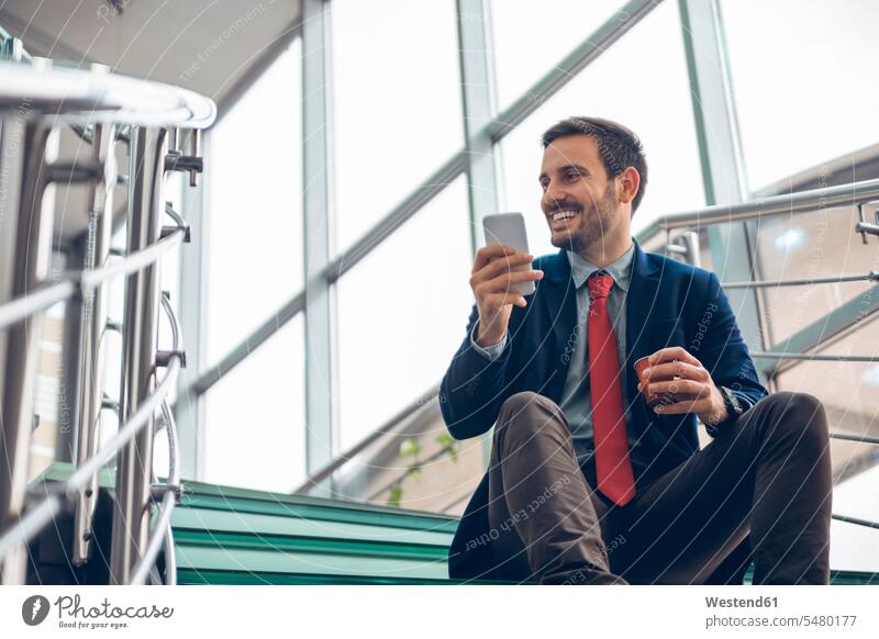 Lächelnder Geschäftsmann sitzt auf der Treppe und benutzt sein Handy Businessmann Businessmänner Geschäftsmänner Smartphone iPhone Smartphones Geschäftsleute
