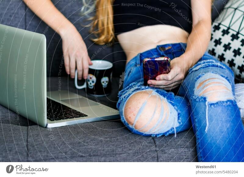 Junge Frau sitzt auf der Couch und benutzt Smartphone und Laptop Gemütlich Wohlfühlen behaglich Gemütlichkeit SMS Textnachricht lesen Lektüre sitzen sitzend