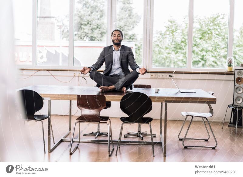 Junger Geschäftsmann sitzt im Schneidersitz auf dem Schreibtisch im Büro Yoga Yoga-Übungen Yogauebungen Yogaübungen Jogauebung Jogauebungen meditieren sitzen
