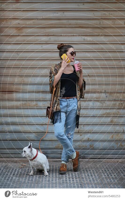 Junge Frau trinkt einen Smoothie und telefoniert mit einem Smartphone, während sie mit ihrem Hund auf der Straße wartet Spanien Haarknoten Dutt Chignons