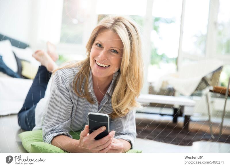 Reife Frau liegt auf dem Boden und benutzt ein Smartphone Wohnzimmer Wohnraum Wohnung Wohnen Wohnräume Wohnungen blond blonde Haare blondes Haar iPhone