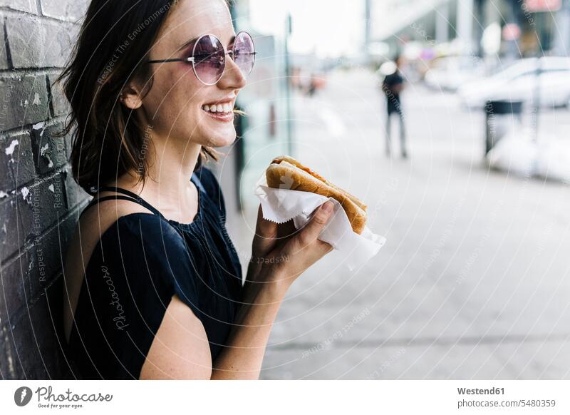 Lächelnde Frau mit an die Wand gelehntem Hot Dog Hot Dogs weiblich Frauen Erwachsener erwachsen Mensch Menschen Leute People Personen essen essend Sonnenbrille