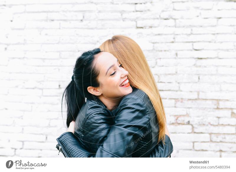 Zwei junge Frauen umarmen sich vor einer weißen Backsteinmauer Freundinnen weiblich Umarmung Umarmungen Arm umlegen Freunde Freundschaft Kameradschaft