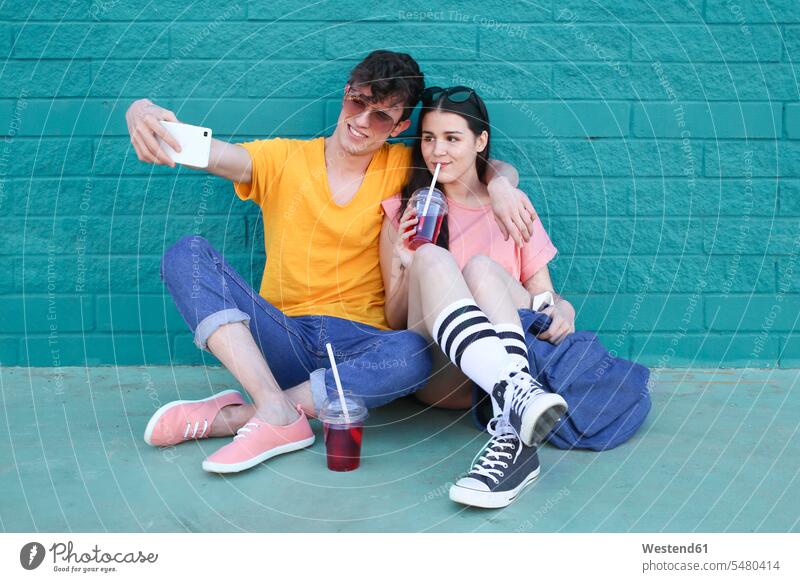 Junges Paar mit alkoholfreien Getränken macht Selfie mit Smartphone vor blauer Backsteinmauer Selfies Pärchen Paare Partnerschaft Mensch Menschen Leute People
