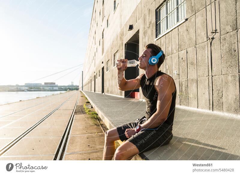 Junger Athlet mit Kopfhörern, der an einer Wand sitzt Pause Pause machen Sportler Musik Kopfhoerer trinken Mauer Mauern sitzen sitzend Jogger Wände Waende