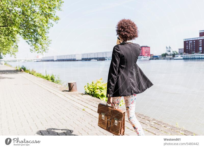 Deutschland, Mannheim, Rückansicht einer jungen Geschäftsfrau mit Aktentasche und Kaffee für einen Spaziergang am Flussufer Geschäftsfrauen Businesswomen