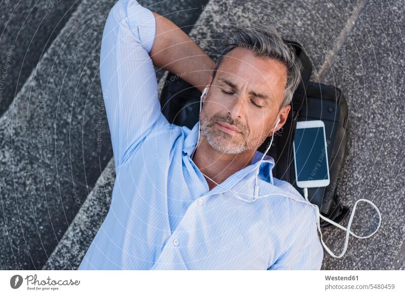 Mann mit geschlossenen Augen, der mit Handy und Ohrstöpseln auf der Treppe liegt Männer männlich entspannt entspanntheit relaxt liegen liegend Mobiltelefon