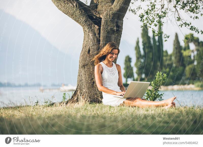 Italien, Gardasee, junge Frau, die sich mit einem Laptop an einen Baum lehnt sitzen sitzend sitzt lächeln Notebook Laptops Notebooks weiblich Frauen Bäume