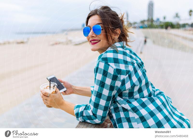 Junge Frau am Strand mit Tasse Kaffee und Smartphone Handy Mobiltelefon Handies Handys Mobiltelefone attraktiv schoen gut aussehend schön Attraktivität