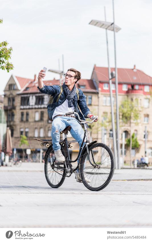 Verspielter junger Mann mit Fahrrad in der Stadt mit dem Handy Selfie Selfies Mobiltelefon Handies Handys Mobiltelefone Männer männlich Bikes Fahrräder Räder