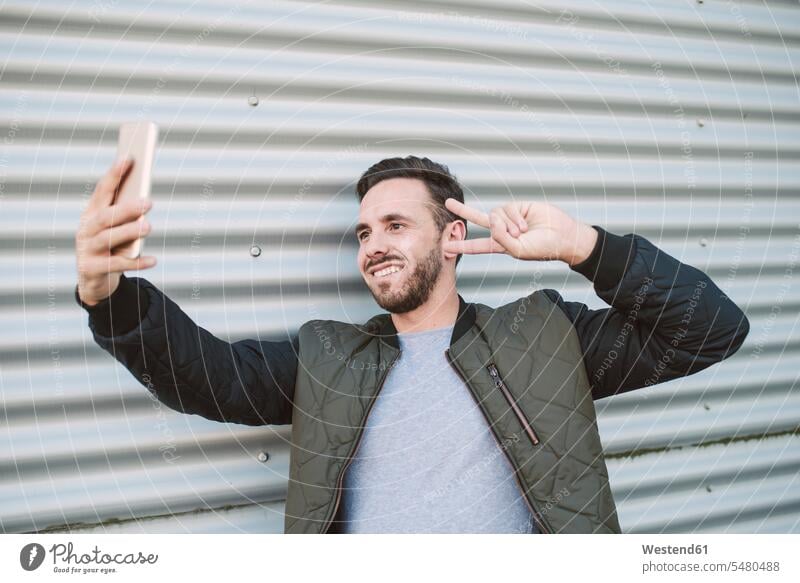 Porträt eines lächelnden Mannes, der ein Selfie mit einem Smartphone macht, das das Siegeszeichen zeigt Victory-Zeichen V Zeichen Victory Zeichen Männer