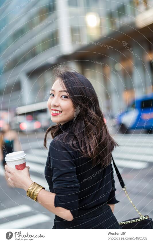 USA, New York City, Manhattan, Porträt einer lächelnden jungen Frau mit Kaffee zum Überqueren der Straße weiblich Frauen Coffee to go zum mitnehmen Portrait