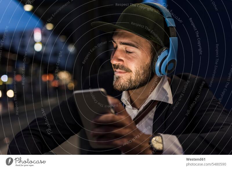 Junger Mann in der Stadt mit Kopfhörern und Handy am Abend Mobiltelefon Handies Handys Mobiltelefone Männer männlich Kopfhoerer staedtisch städtisch lächeln