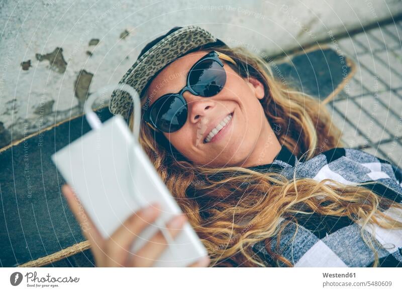 Lächelnde junge Frau liegt auf Skateboard und schaut auf Handy weiblich Frauen Mobiltelefon Handies Handys Mobiltelefone liegen liegend lächeln Erwachsener