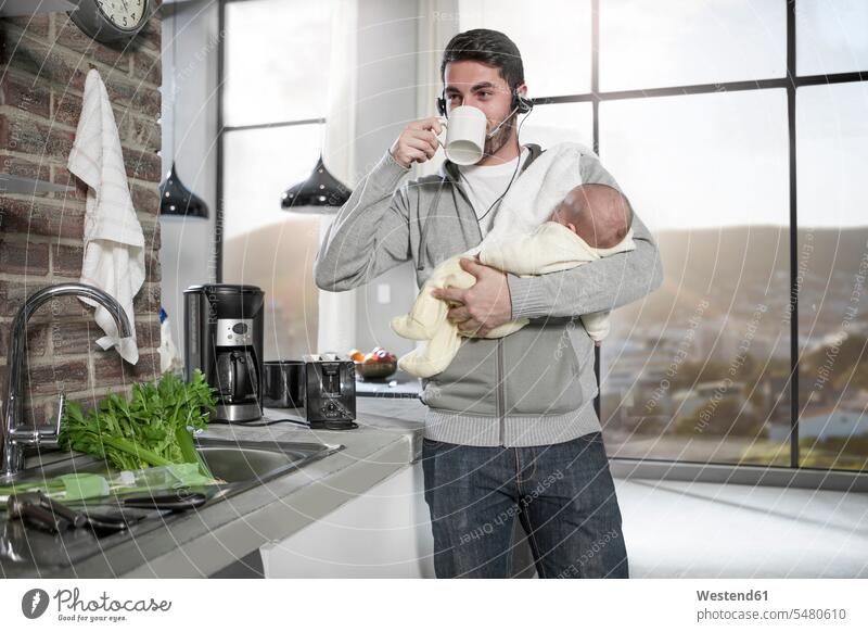 Vater mit Headset trinkt Kaffee in der Küche und hält Baby telefonieren anrufen Anruf telephonieren Papas Väter Vati Vatis Papis Küchen trinken Telefon benutzen