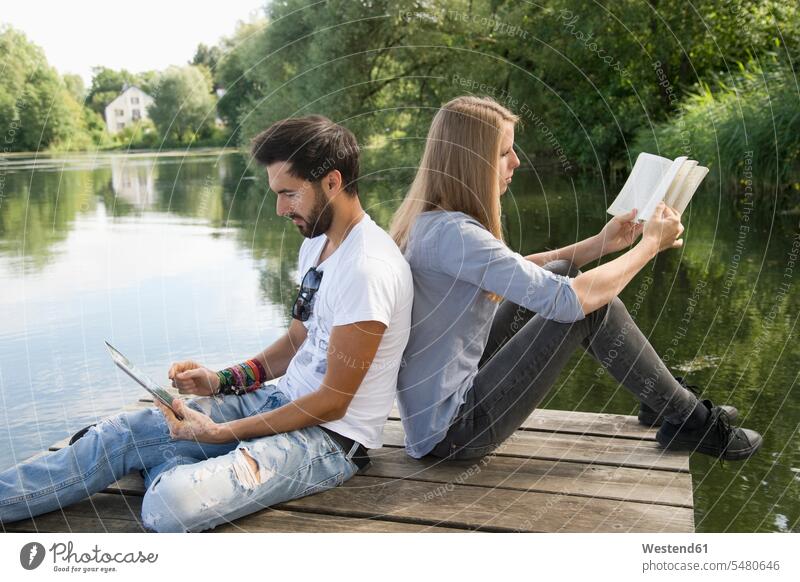 Junges Paar sitzt mit Buch und Tafel auf einem Steg an einem See Tablet Tablet Computer Tablet-PC Tablet PC iPad Tablet-Computer lesen Lektüre Bücher Stege