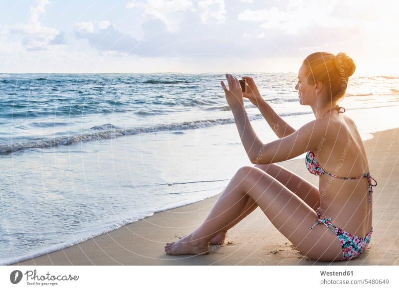 Dominikanische Republik, Junge Frau am tropischen Strand beim Fotografieren mit mobilem Gerät sitzen sitzend sitzt weiblich Frauen Beach Straende Strände