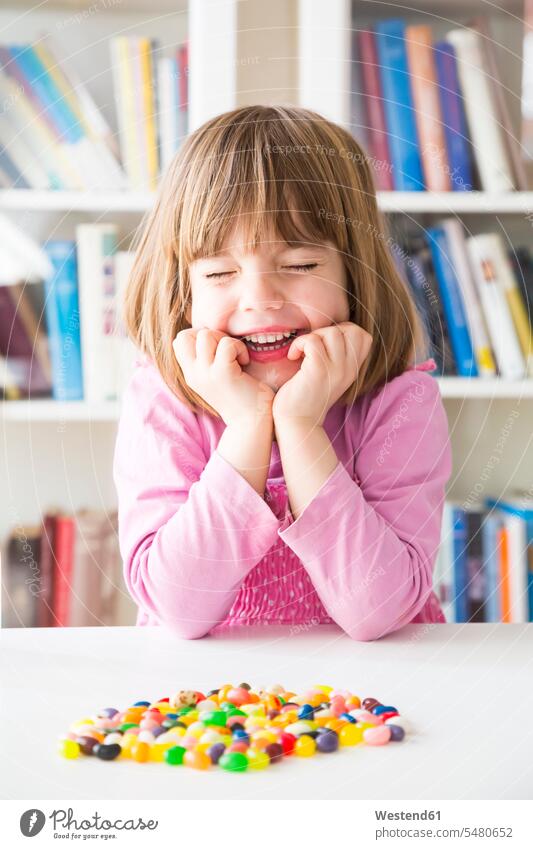 Bildnis eines lächelnden kleinen Mädchens mit geschlossenen Augen mit Geleebohnen auf einem Tisch liegend Bücherregal Bücherregale erwartungsvoll Erwartung