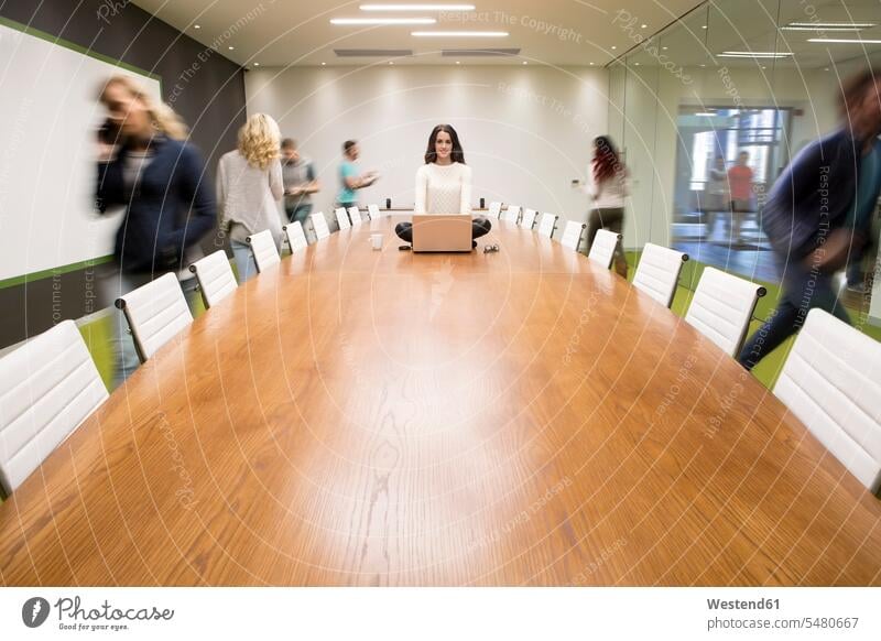Geschäftsfrau mit Laptop, die auf einem Konferenztisch sitzt, auf dem Leute herumlaufen entspannt entspanntheit relaxt Kollegin Kolleginnen Büro Office Büros