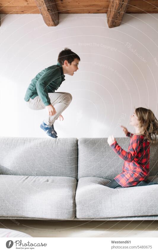 Junge springt auf der Couch in die Luft, während seine Schwester ihn beobachtet Sofa Couches Liege Sofas Schwestern zuschauen beobachten anschauen betrachten