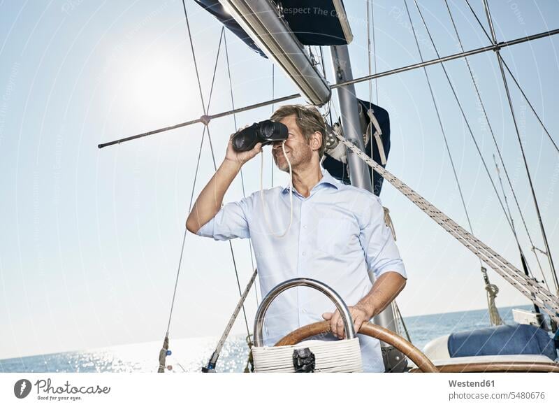 Erwachsener Mann benutzt Fernglas am Steuer seines Segelbootes Männer männlich Ferngläser Feldstecher Fernglaeser Segeln segelnd segelt erwachsen Mensch