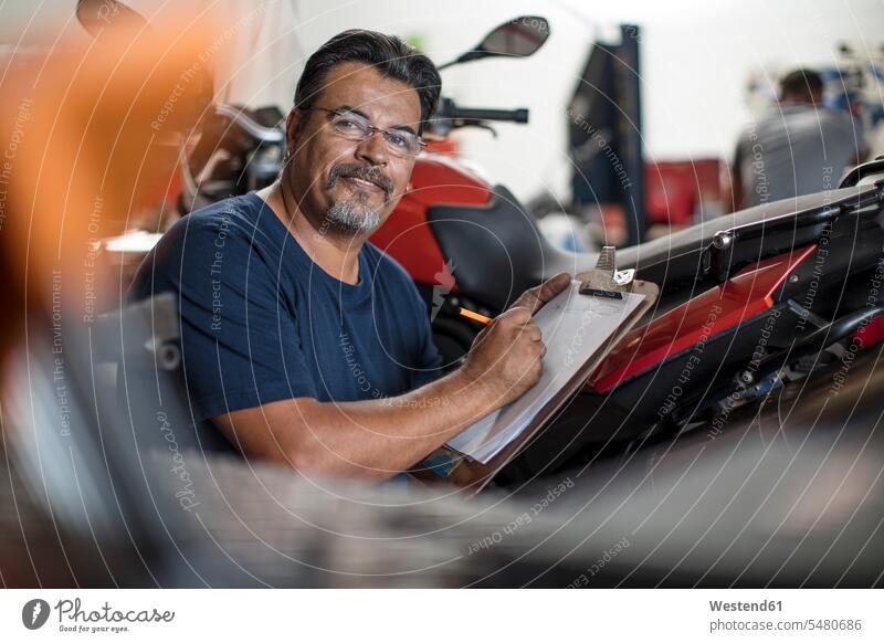 Porträt eines lächelnden Mechanikers mit Klemmbrett in der Motorradwerkstatt reparieren Reparatur Monteur Motorräder Klemmbretter arbeiten Arbeit Industrie