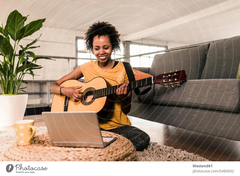 Lächelnde junge Frau zu Hause, die Gitarre spielt und auf den Laptop schaut Notebook Laptops Notebooks lächeln weiblich Frauen Gitarren Computer Rechner