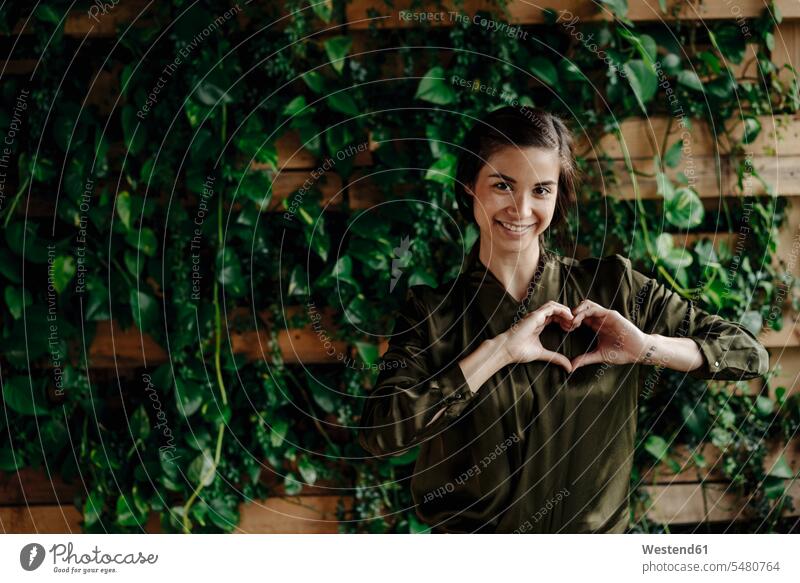 Porträt einer lächelnden jungen Frau, die ihr Herz an einer Wand mit Kletterpflanzen formt Umwelt Ökologie weiblich Frauen Portrait Porträts Portraits Herzform