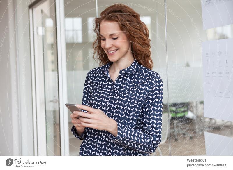 Lächelnde Frau im Büro schaut auf Handy weiblich Frauen lächeln Office Büros Mobiltelefon Handies Handys Mobiltelefone Erwachsener erwachsen Mensch Menschen