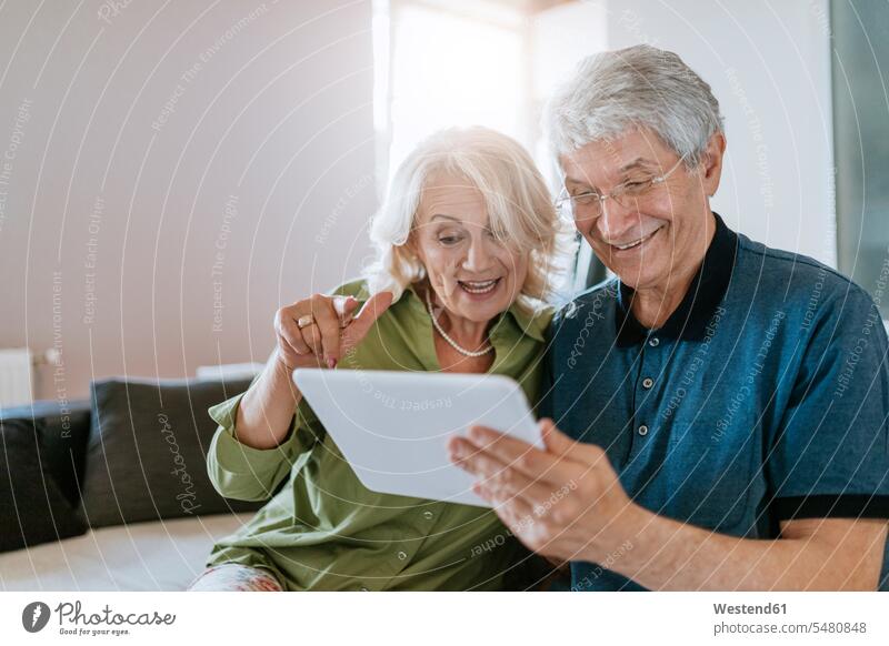 Glückliches älteres Ehepaar sitzt zu Hause auf der Couch und teilt sich ein Tablet Paar Pärchen Paare Partnerschaft lachen Sofa Couches Liege Sofas