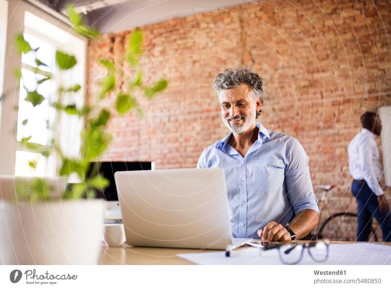Lächelnder Geschäftsmann mit Laptop im Büro mit Kollege im Hintergrund lächeln Office Büros Notebook Laptops Notebooks Businessmann Businessmänner