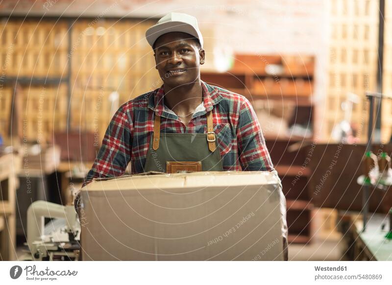 Porträt eines lächelnden Mannes, der einen Karton in der Werkstatt trägt Männer männlich arbeiten Arbeit Pappkartons Kartons Werkstätte Werkstaette Werkstaetten