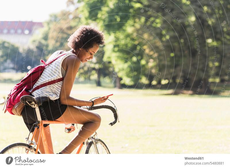 Junge Frau auf Fahrrad im Park beim Handy-Check Bikes Fahrräder Räder Rad lächeln Parkanlagen Parks weiblich Frauen Mobiltelefon Handies Handys Mobiltelefone