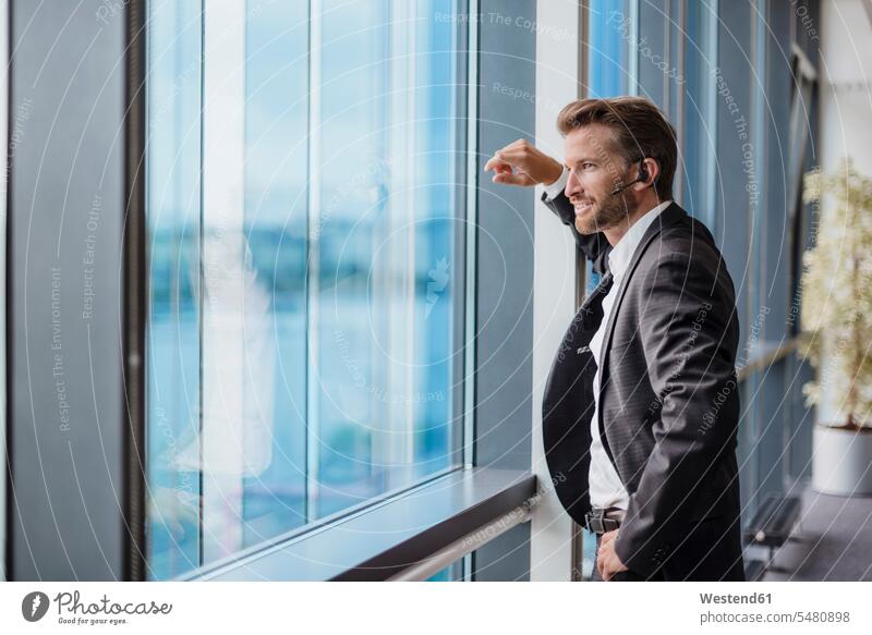 Entspannter Geschäftsmann mit Bluetooth-Headset steht in seinem Büro und schaut aus dem Fenster Headsets telefonieren anrufen Anruf telephonieren Businessmann